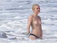 She Sits Nude on the Sand Between Ocean Waves and Cliffs