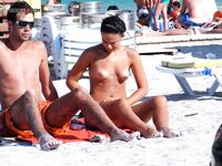 Two Nudists Stand on Sandy Shore With Shorts and Sunscreen Nearby