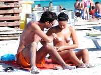 Two Nudists Stand on Sandy Shore With Shorts and Sunscreen Nearby