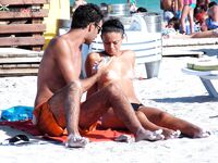 Two Nudists Stand on Sandy Shore With Shorts and Sunscreen Nearby