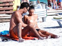 Two Nudists Stand on Sandy Shore With Shorts and Sunscreen Nearby