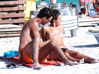 Two Nudists Stand on Sandy Shore With Shorts and Sunscreen Nearby