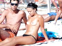 Two Nudists Stand on Sandy Shore With Shorts and Sunscreen Nearby